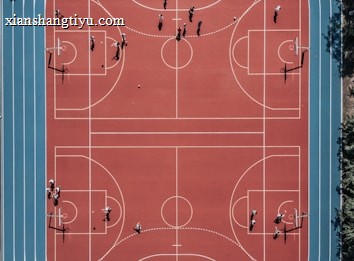 An aerial view of a large, vibrant sports complex featuring a red basketball court with multiple hoops and a surrounding blue running track. Several people are scattered across the area, engaged in various activities.