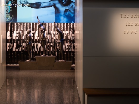 A dimly lit display features statues of athletes standing on a podium. They are positioned in front of a backdrop with the words 'LEVELING THE PLAYING FIELD' in bold letters. On the wall to the right, a softly lit quote is partially visible. The overall ambiance suggests a museum or exhibition setting.