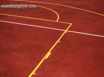 A sports court with a reddish-brown surface and bright yellow and white lines. The lines form geometric shapes, including rectangles and circles, suggesting markings for games like basketball or netball.