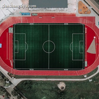 An aerial view of a large sports complex featuring a central green football field surrounded by a red running track. Adjacent to the track, there is a parking area, buildings, and pathways. Nearby, there are additional sports courts in different colors, including a blue swimming pool and orange basketball courts.