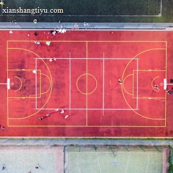 An aerial view of a sports court with a vibrant red surface and yellow lines marking different sections for basketball and soccer. Several people are present on the court, some engaged in activities, while others are standing along the perimeter. The court is enclosed by a metal fence and surrounded by grass on all sides.