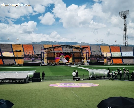A sports stadium with a large stage set up on the field, hosting an outdoor event. Spectators wearing colorful hats sit in the foreground. The seating areas display vibrant colors, and a clear blue sky with scattered clouds is visible. Large screens display graphics related to the event.