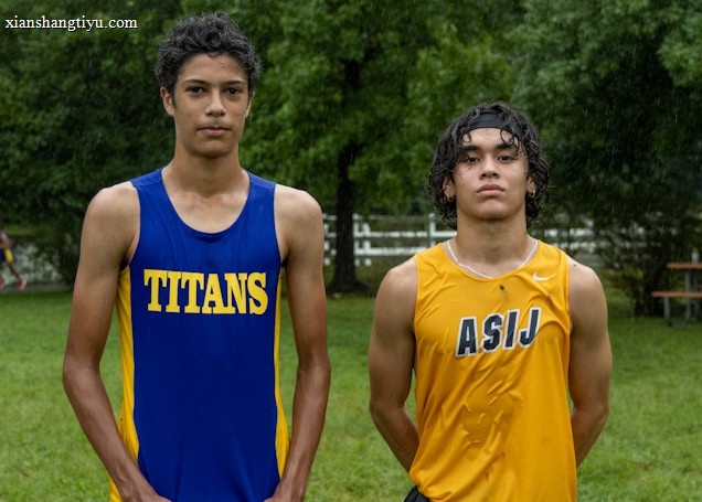 Two young athletes wearing sleeveless sports tops stand on a grassy field. The athlete on the left is wearing a blue top with the word 'TITANS' while the one on the right wears a yellow top with 'ASIJ'. The background features green trees and a white fence, with a bench visible on the right.