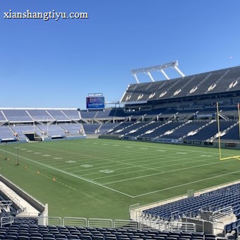 A large, empty sports stadium with blue seats surrounding a well-maintained green grass field. Bright sunlight creates clear shadows, and there are two goal posts on opposite ends of the field. A big electronic scoreboard displays some graphics, and the sky is clear and blue.
